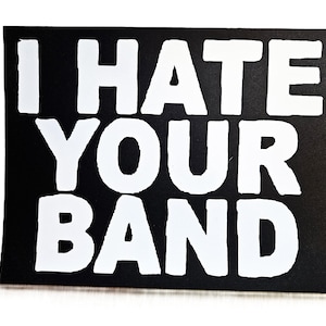 Puede incluir: Una pegatina rectangular negra con el texto blanco "I HATE YOUR BAND". El texto está en negrita, con una fuente sans-serif. La pegatina está sobre un fondo blanco.