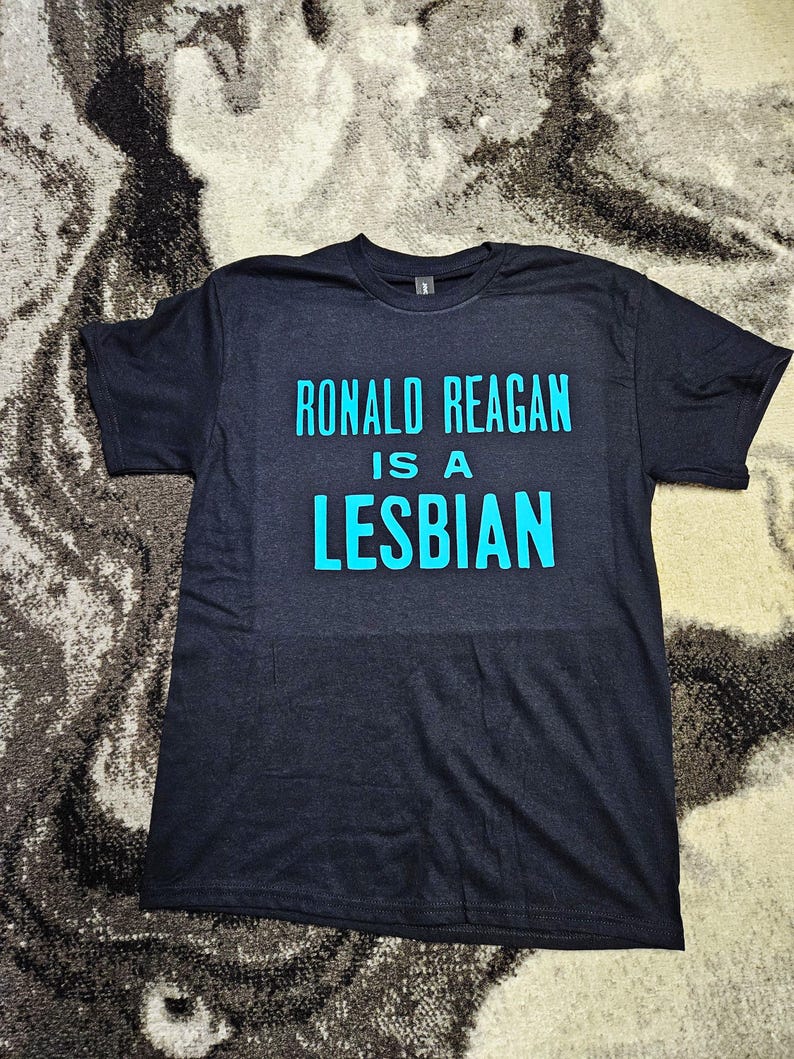 May include: A black short-sleeved t-shirt featuring the text "RONALD REAGAN IS A LESBIAN" printed in a bright cyan blue font across the chest. The shirt has a crew neckline and a standard fit, with the text centered in a bold, sans-serif typeface.