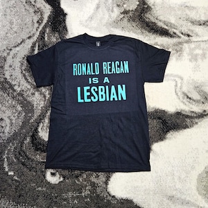 May include: A black short-sleeved t-shirt featuring the text RONALD REAGAN IS A LESBIAN printed in a bold, light blue sans-serif font across the chest. The garment has a crew neckline and a standard fit, displaying clear, legible lettering.