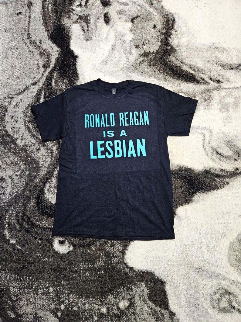 May include: A black short-sleeved t-shirt featuring the text RONALD REAGAN IS A LESBIAN printed in a bold, light blue sans-serif font across the chest. The garment has a crew neckline and a standard fit, displaying clear, legible lettering.