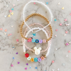 May include: Four bracelets with colorful beads spelling out "DISNEY" and "MAGIC" with a white Mickey Mouse charm. The bracelets are on a marble surface with scattered Mickey Mouse confetti.