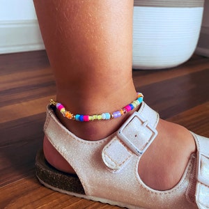 May include: A colorful beaded anklet with a gold clasp. The anklet is made with small, round beads in shades of pink, blue, green, yellow, and orange. The anklet is worn on a person's ankle.