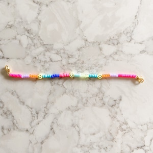 May include: A colourful beaded bracelet with three gold smiley face charms. The bracelet is made with pink, orange, yellow, green, blue, and purple beads.