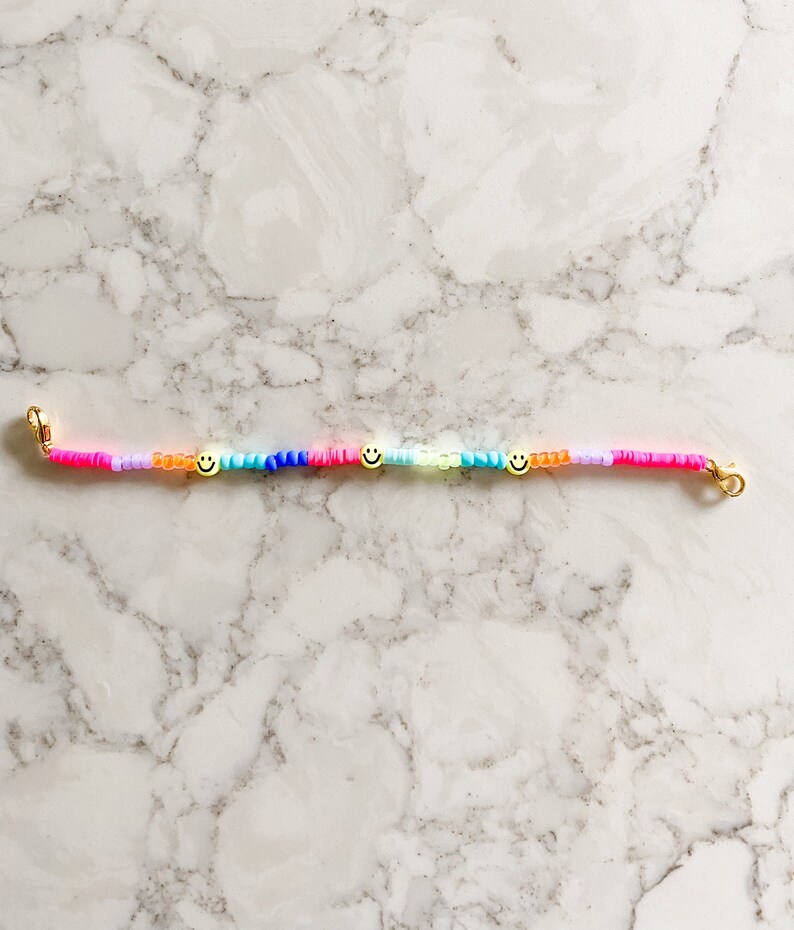 May include: A colourful beaded bracelet with three gold smiley face charms. The bracelet is made with pink, orange, yellow, green, blue, and purple beads.