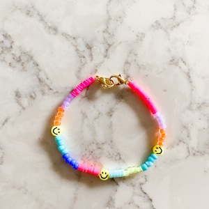 May include: A rainbow-coloured beaded bracelet with three yellow smiley face charms. The bracelet is made with small, round beads in shades of pink, purple, blue, green, and yellow.