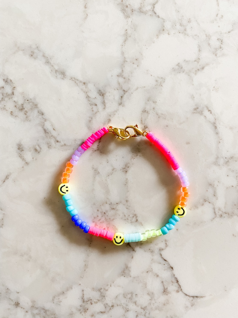 May include: A rainbow-coloured beaded bracelet with three yellow smiley face charms. The bracelet is made with small, round beads in shades of pink, purple, blue, green, and yellow.