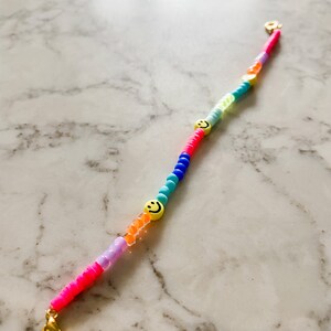May include: A colourful beaded bracelet with a gold clasp and two yellow smiley face charms. The beads are pink, purple, orange, blue, and green.