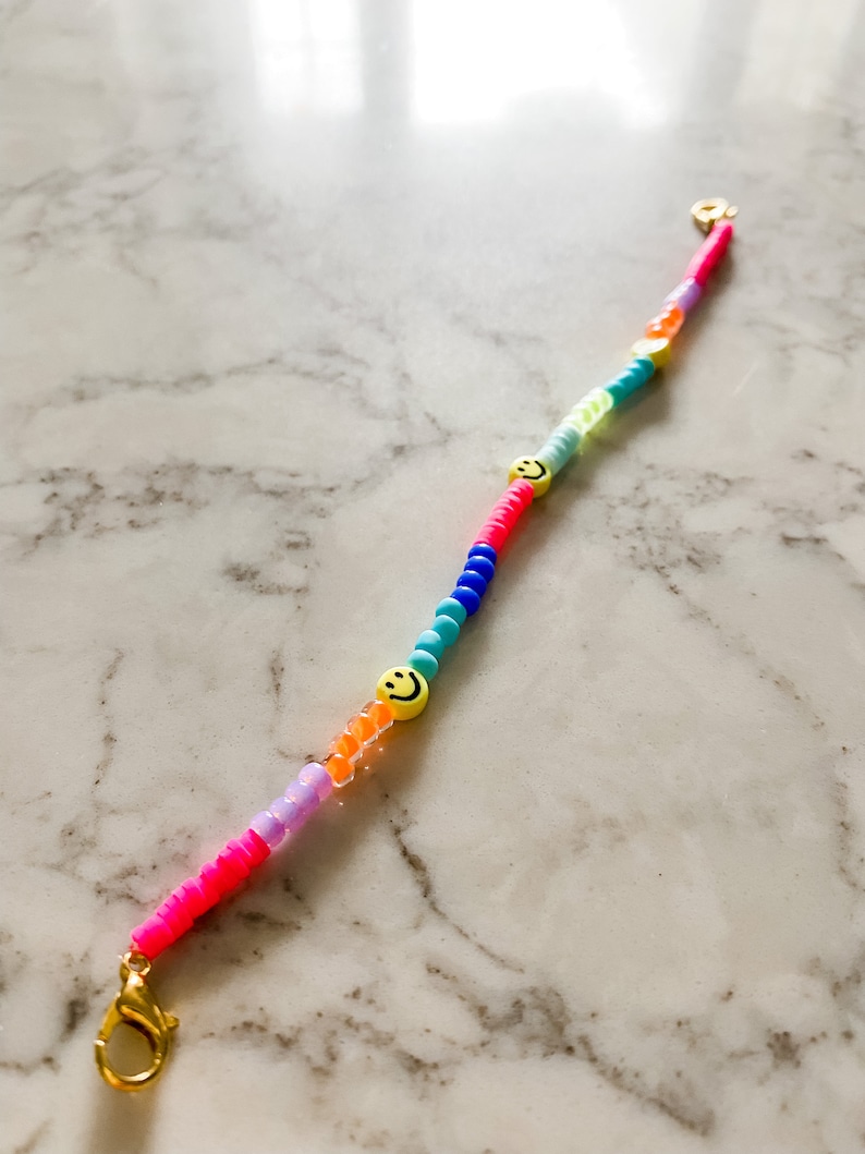 May include: A colourful beaded bracelet with a gold clasp and two yellow smiley face charms. The beads are pink, purple, orange, blue, and green.