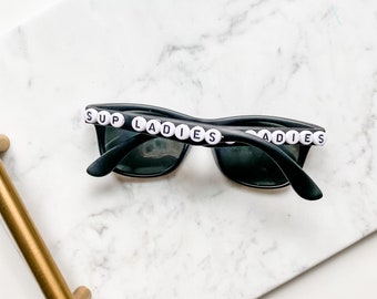 May include: Black sunglasses with white beads spelling "SUP LADIES" on the top of the frames.