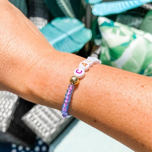 May include: A white beaded bracelet with the word "CAL" spelled out in colorful letters. The bracelet is on a person's wrist and has a gold bead in the center.