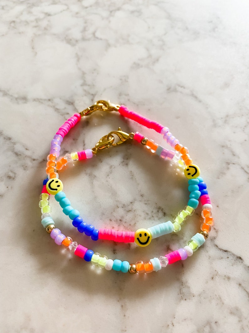 May include: Two colourful beaded bracelets with smiley face charms. The bracelets are made with pink, blue, orange, yellow, green and white beads.