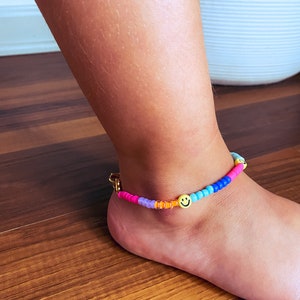 May include: A colourful beaded anklet with a gold clasp and a smiley face charm. The anklet is made with pink, purple, blue, orange, and yellow beads.