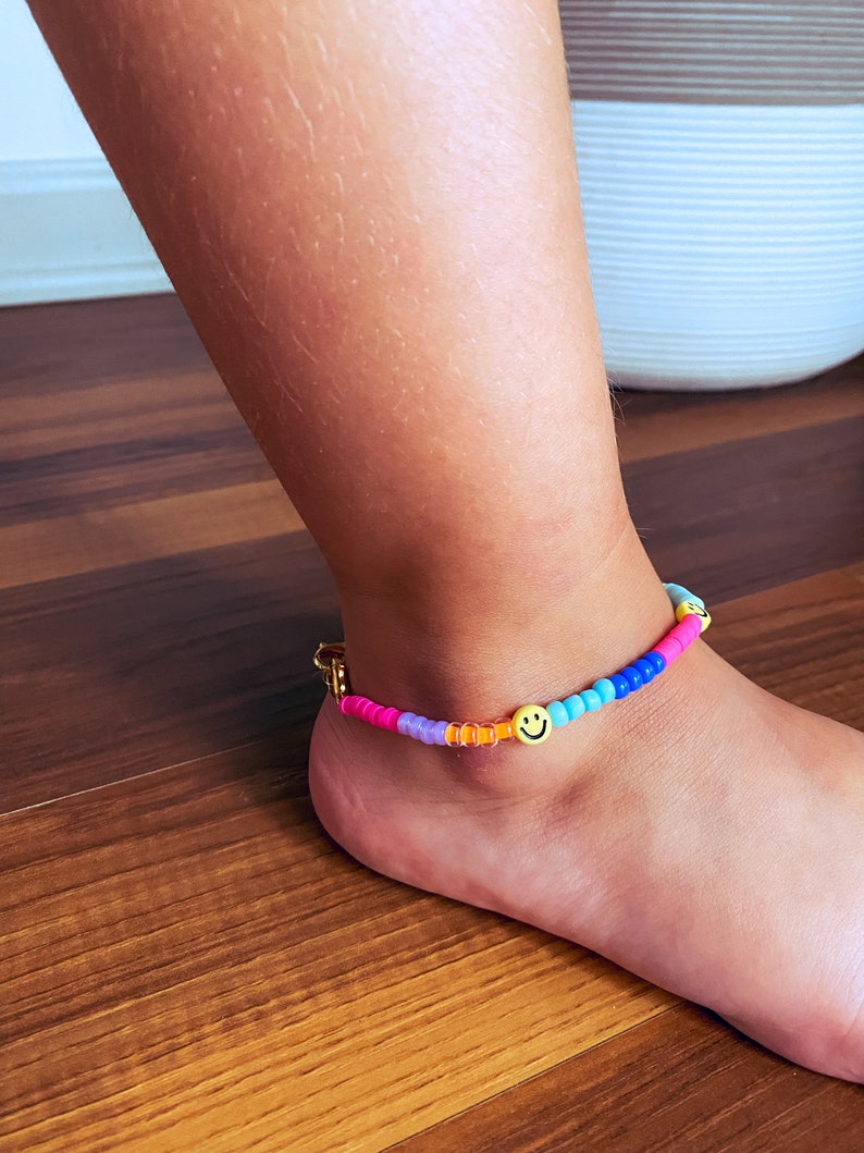 May include: A colourful beaded anklet with a gold clasp and a smiley face charm. The anklet is made with pink, purple, blue, orange, and yellow beads.