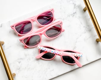May include: Three pairs of pink sunglasses with dark lenses rest on a white marble tray with gold handles. One pair of sunglasses has the words "VACAY MODE" spelled out in colorful letter beads across the frame.