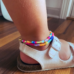 May include: A colourful beaded anklet with a yellow smiley face charm. The anklet is worn on a person's ankle with white sandals.