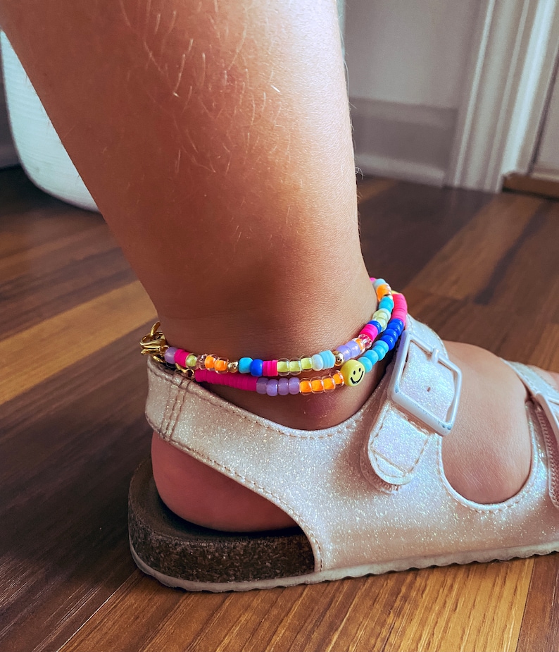 May include: A colourful beaded anklet with a yellow smiley face charm. The anklet is worn on a person's ankle with white sandals.