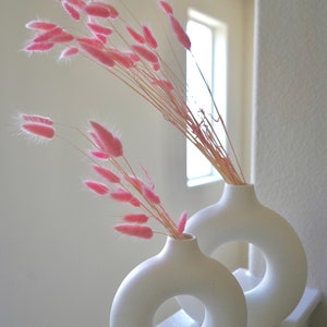 May include: Two white ceramic vases with a donut-shaped opening. The vases are filled with pink dried pampas grass.