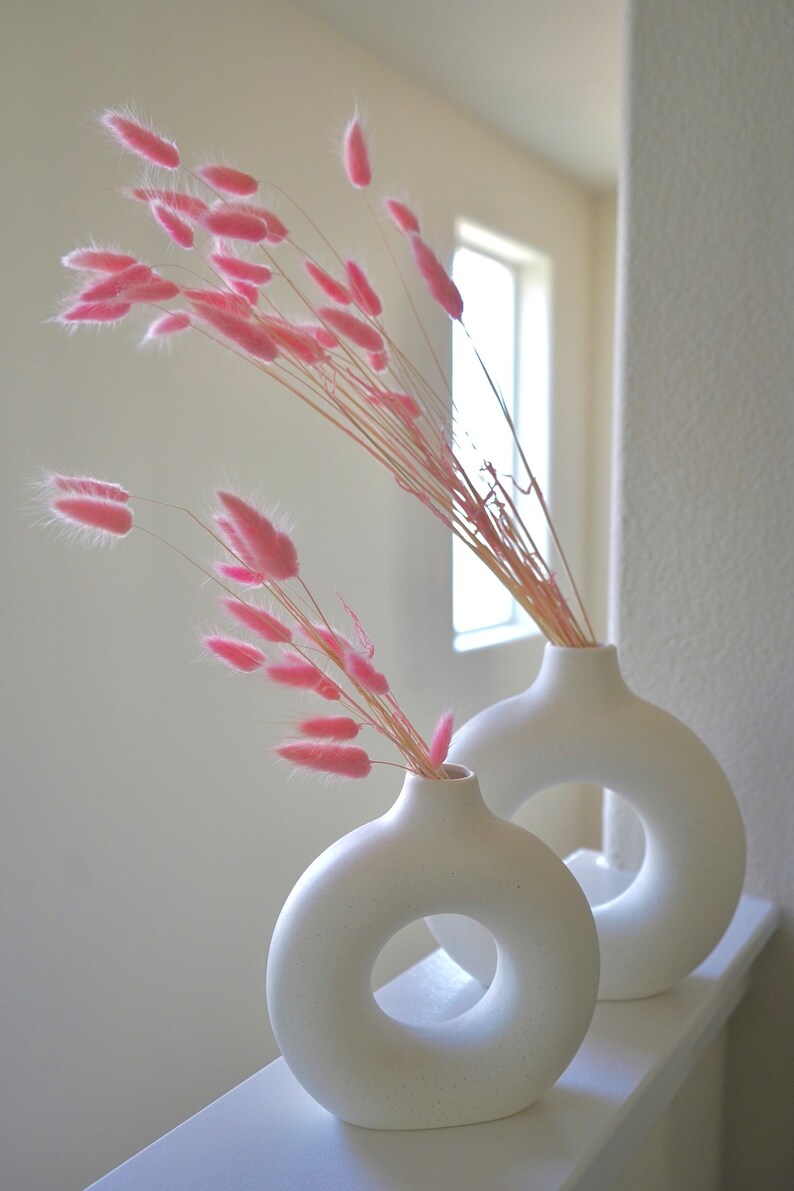 May include: Two white ceramic vases with a donut-shaped opening. The vases are filled with pink dried pampas grass.