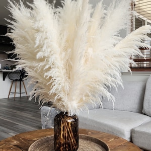 May include: A large arrangement of fluffy, cream-colored pampas grass in a dark brown, speckled glass vase. The vase sits on a round, woven tray atop a wooden coffee table. The background includes a gray sofa and a window.