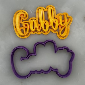 以下が含まれることがあります： 「Gabby」という名前の形をした金色のプラスチック製のクッキーカッターです。