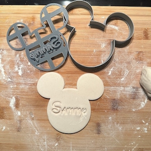 May include: A close-up of a wooden surface with two grey plastic cookie cutters shaped like Mickey Mouse heads. One cutter has the name "Sammy" inscribed on it. A piece of dough is also visible on the surface.