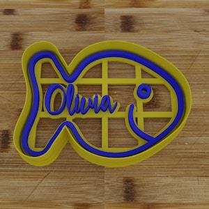 Puede incluir: Un cortador de galletas de plástico amarillo y azul con forma de pez con el nombre "Olivia" escrito en su interior.