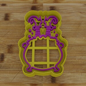 May include: A yellow and purple plastic cookie cutter shaped like a cauldron with a lid. The cauldron has a grid pattern on the front and a decorative design on the lid.