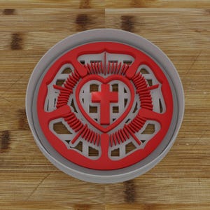 May include: A red and white cookie cutter featuring a heart with a cross inside, surrounded by a stylized rose design. The cutter is round and sits inside a gray container, set against a wooden background.