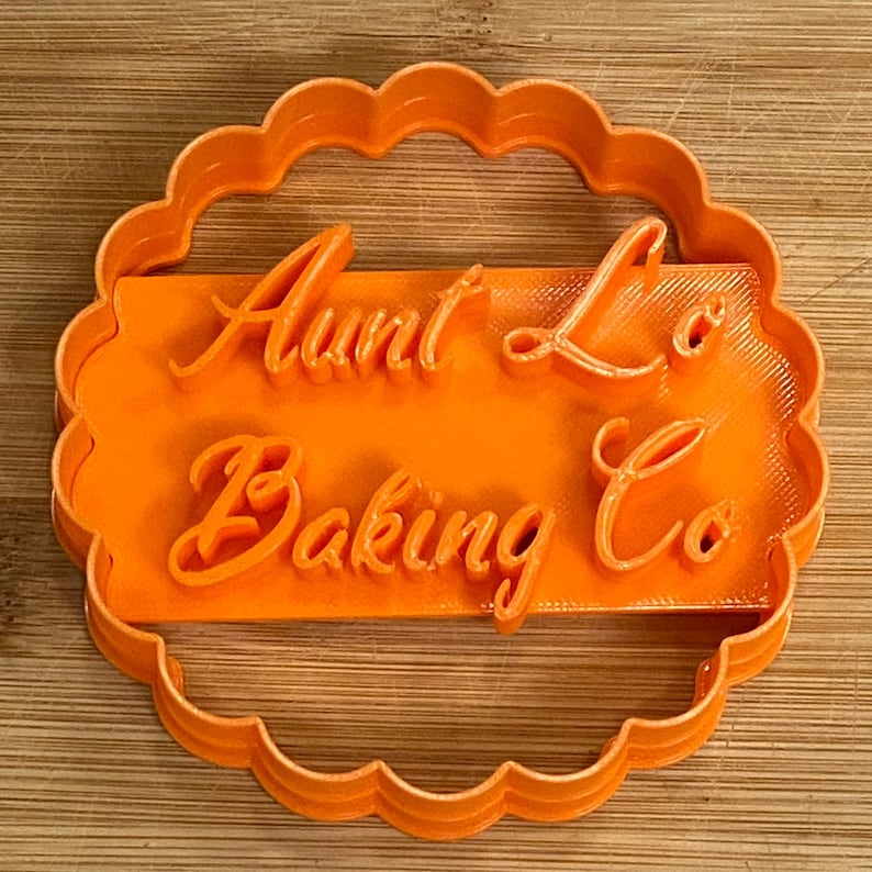 Puede incluir: Cortador de galletas de pl&aacute;stico naranja con borde festoneado. El cortador tiene forma de c&iacute;rculo con un recorte rectangular en el centro. El texto "Aunt Lo's Baking Co" est&aacute; escrito en una fuente cursiva dentro del recorte.