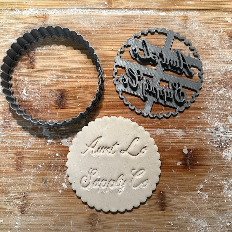 Puede incluir: Primer plano de un cortador de galletas y un c&iacute;rculo de masa para galletas. El cortador de galletas es un c&iacute;rculo con las palabras "Aunt Lo Supply Co" recortadas. El c&iacute;rculo de masa para galletas tiene las palabras "Aunt Lo Supply Co" impresas.