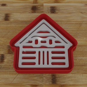 May include: Red plastic cookie cutter shaped like a house with a white outline. The house has a roof with a decorative design and a series of horizontal lines on the front.