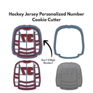May include: A set of hockey jersey-shaped cookie cutters. The cutters are gray and red, with the text "Hockey Jersey Personalized Number Cookie Cutter" at the top. The image also shows a gray cookie with the number 37.