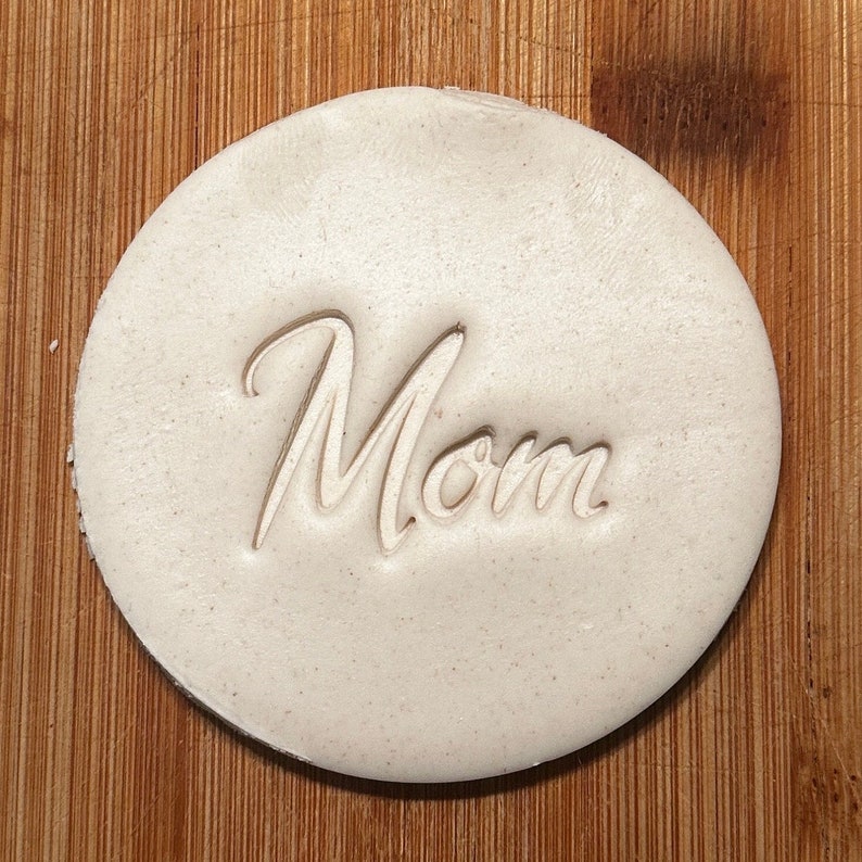 Puede incluir: Un adorno redondo de arcilla blanca con la palabra "Mam&aacute;" en relieve en una fuente cursiva.