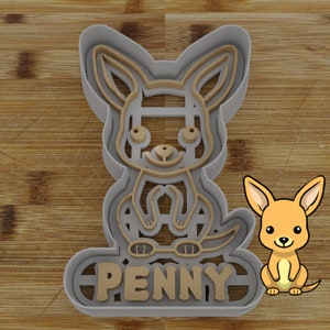 May include: A gray and tan kangaroo-shaped cookie cutter with the name "PENNY" at the bottom. A cartoon kangaroo illustration is next to the cookie cutter. The cookie cutter is on a wooden surface.
