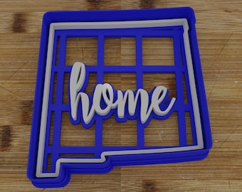 May include: A blue and white cookie cutter in the shape of New Mexico with the word "home" in white script. The cutter has a grid design within the state outline, and sits on a wooden surface.