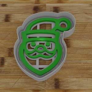May include: A green plastic cookie cutter shaped like a Santa Claus face. The cutter has a detailed design with a hat, beard, and eyes.