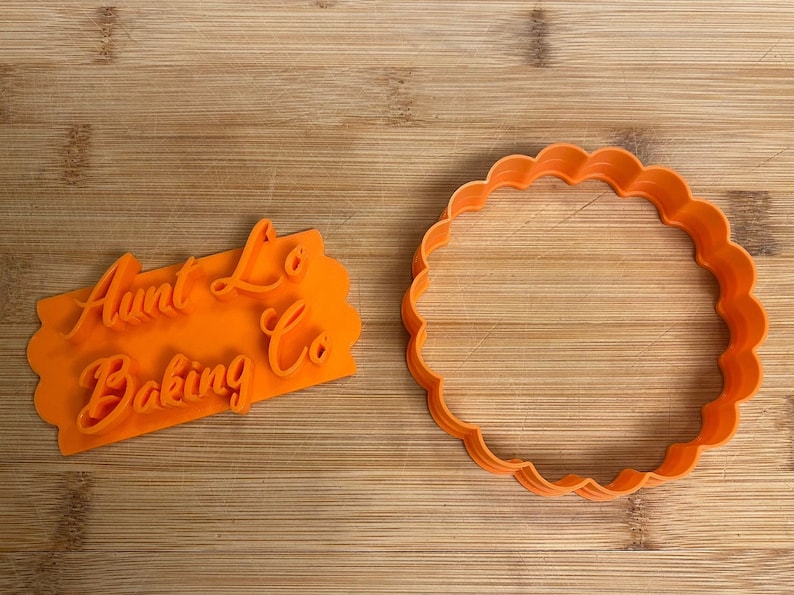 Puede incluir: Dos cortadores de galletas de pl&aacute;stico naranja. Uno tiene forma de c&iacute;rculo con un borde festoneado. El otro es rectangular con las palabras "Aunt Lo's Baking Co" en una fuente cursiva.