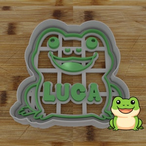 May include: A gray plastic cookie cutter shaped like a frog with the name Luca written inside. A cartoon frog is sitting next to the cutter.
