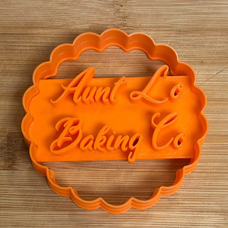 Puede incluir: Cortador de galletas de pl&aacute;stico naranja en forma de c&iacute;rculo con un borde festoneado. El cortador tiene las palabras "Aunt Lo's Baking Co" escritas en una fuente cursiva.