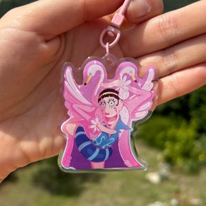 Peut inclure: Un personnage rose et blanc avec de grands yeux et une coiffe à plumes rose et blanche. Le personnage tient une fleur rose dans chaque main. Le personnage est entouré de deux flamants roses. Le personnage est sur un porte-clés en acrylique transparent.