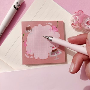 Può includere: Un blocco note rosa con un design a forma di nuvola e linee a griglia, decorato con illustrazioni di dessert. Sono visibili anche una penna bianca con una faccia di panda e una penna rosa. Il blocco note è su carta a righe e una superficie rosa.