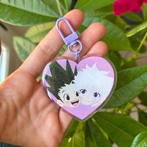 Op de afbeelding: Een roze hartvormige sleutelhanger met een glitterachtergrond waarop twee anime-personages te zien zijn, een met groen haar en de andere met wit haar. De sleutelhanger heeft een paarse metalen sluiting.