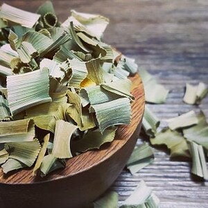 May include: A wooden bowl filled with dried, light green and brown plant leaves. The leaves are scattered on a wooden surface.