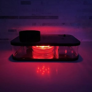 May include: A black rectangular platform with a clear acrylic enclosure. The platform has a black lid and a red LED light inside. The light is reflected on the surface below.