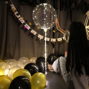 Pode incluir: Um bal&atilde;o transparente com luzes LED brancas dentro, uma corda de luzes e uma faixa que diz "Feliz anivers&aacute;rio". Tamb&eacute;m h&aacute; bal&otilde;es dourados e pretos no ch&atilde;o.
