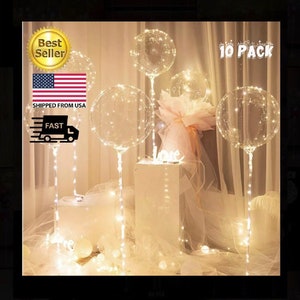May include: Ten clear plastic balloons with white LED lights inside. The balloons are tied to white strings and are arranged in a decorative setting. The text "10 PACK" is visible in the upper right corner of the image.