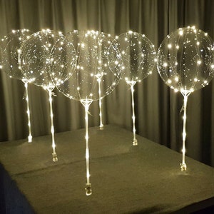 Pode incluir: Um conjunto de seis bal&otilde;es de pl&aacute;stico transparente com luzes LED embutidas. Os bal&otilde;es s&atilde;o iluminados com luzes brancas quentes e est&atilde;o dispostos em fila sobre uma mesa.
