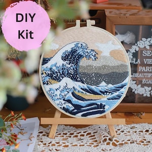 May include: DIY embroidery kit featuring a blue and white wave design in a wooden embroidery hoop. The hoop is on a small wooden easel.