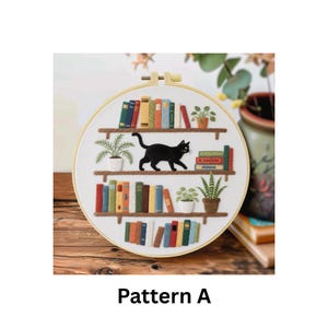 Puede incluir: Arte de aro bordado que representa un gato negro caminando sobre una estanter&iacute;a con libros coloridos y plantas en macetas. El texto "Pattern A" est&aacute; en la parte inferior de la imagen. El aro est&aacute; sobre una superficie de madera.