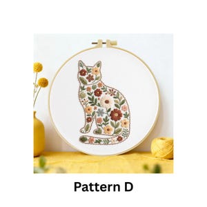 Puede incluir: Un dise&ntilde;o de gato bordado con flores sobre tela blanca, dentro de un bastidor de madera. El gato est&aacute; relleno de flores marrones, naranjas y verdes. El texto "Pattern D" est&aacute; en la parte inferior.